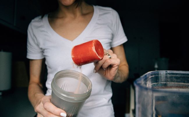 Frau die sich einen Löffel Proteinpulver in ihren Shaker kippt